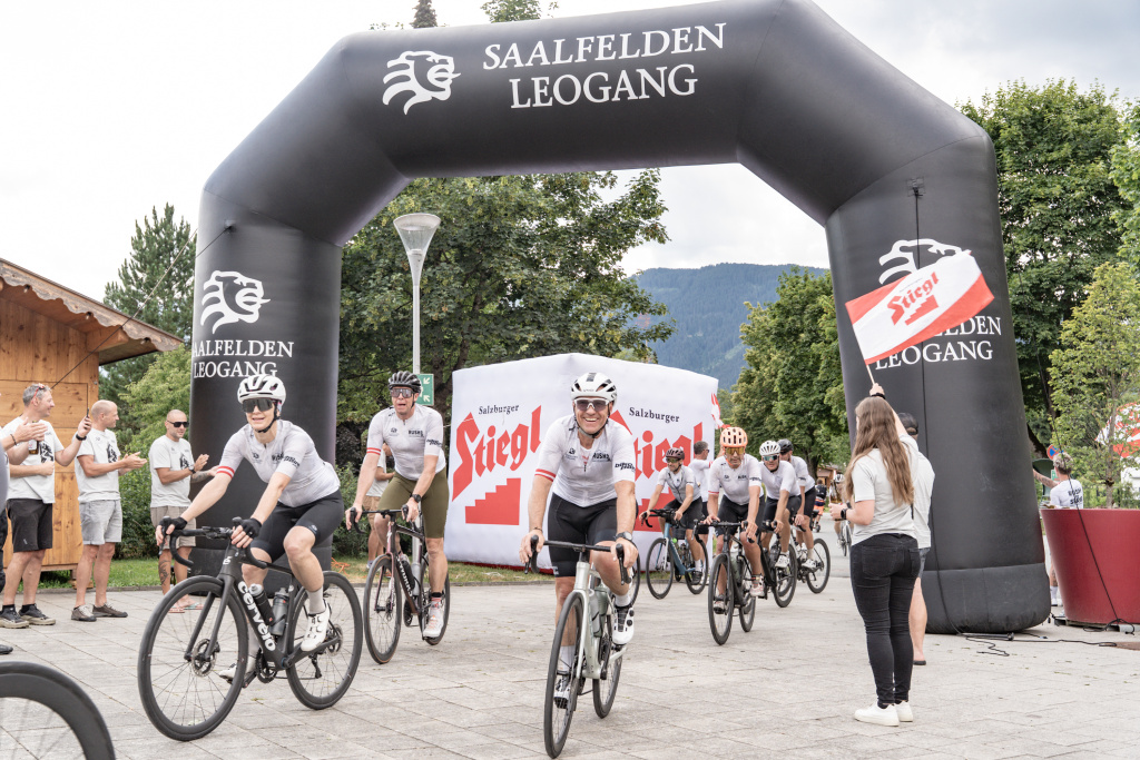 Wielrenners rijden onder een zwarte Saalfelden-Leogang-boog door, toeschouwers klappen